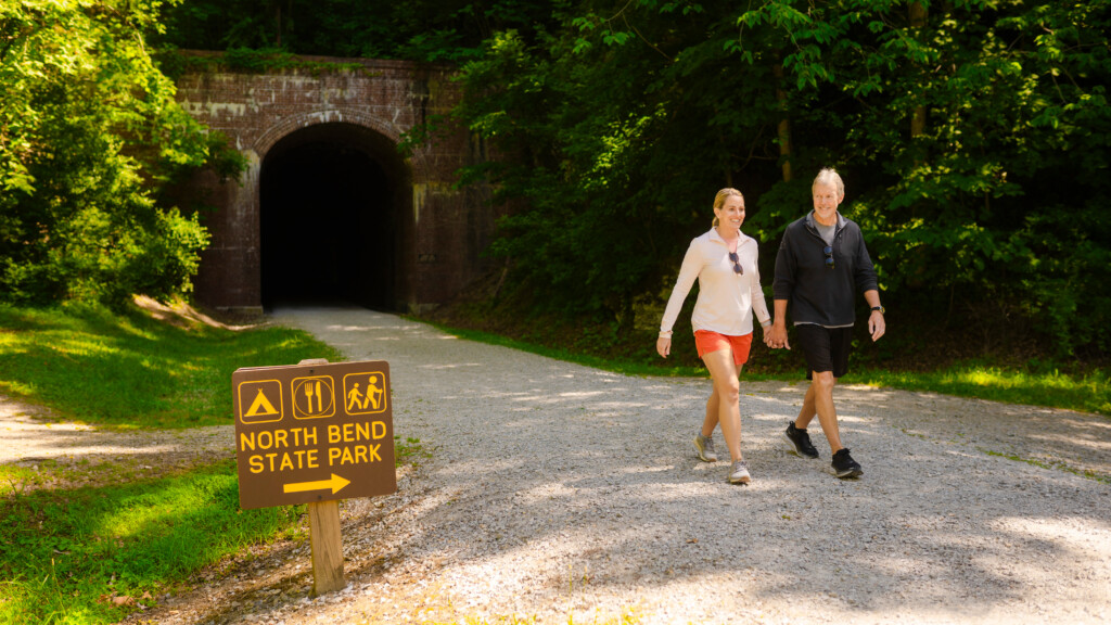 A smiling couple walks hand-in-hand along a gravel path near a historic railroad tunnel at North Bend State Park, surrounded by lush green trees. A brown sign with yellow icons indicates amenities like camping, dining, and hiking.