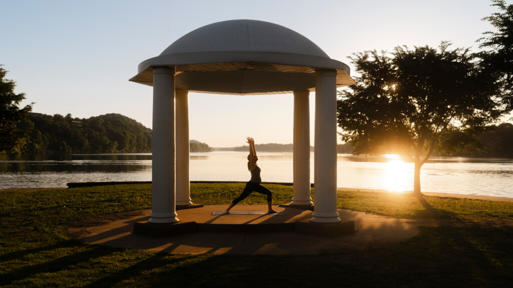 A woman practices yoga at Point Park sunrise beneath a white domed pavilion overlooking a calm Ohio and Little Kanawha rivers, with golden light streaming through nearby trees and reflecting off the water.