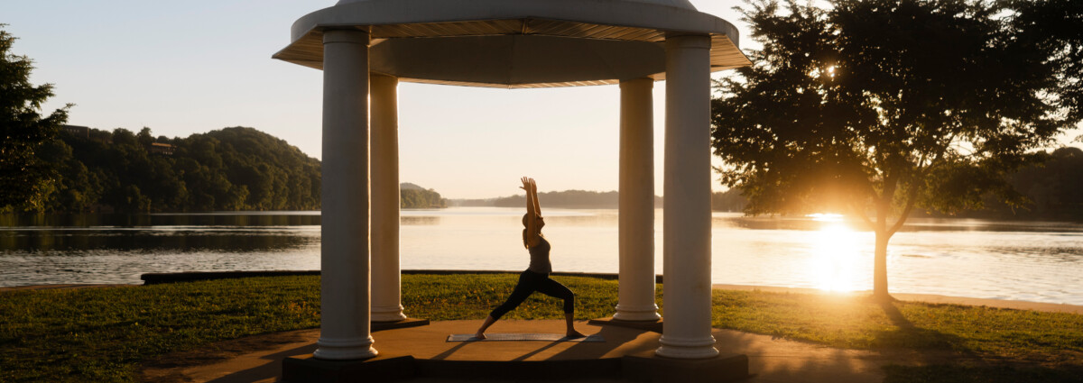 A woman practices yoga at Point Park sunrise beneath a white domed pavilion overlooking a calm Ohio and Little Kanawha rivers, with golden light streaming through nearby trees and reflecting off the water.