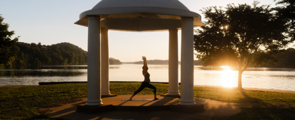 A woman practices yoga at Point Park sunrise beneath a white domed pavilion overlooking a calm Ohio and Little Kanawha rivers, with golden light streaming through nearby trees and reflecting off the water.